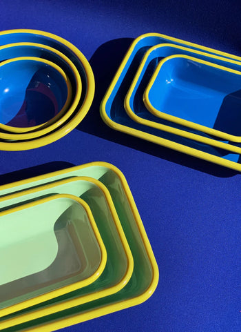 The Bornn Enamelware Colorama Baking Dish, Medium, features a handmade design in blue and green with yellow rims, arranged on a bright blue surface to highlight the brand’s vibrant style.