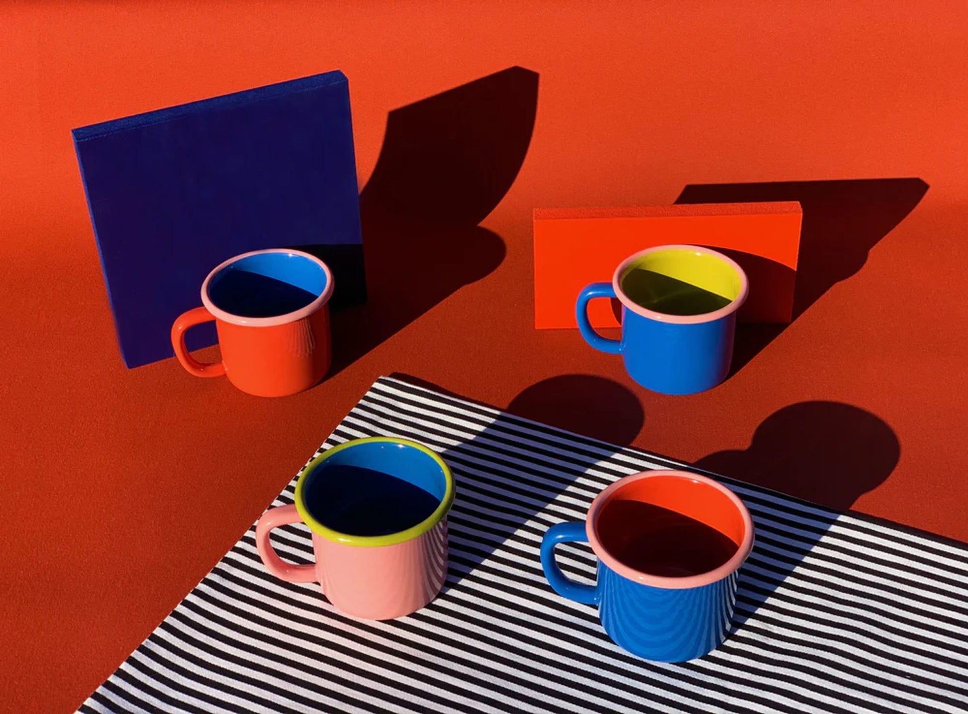 Four Bornn Enamelware Colorama Mugs in red, pink, blue, and orange sit on a red surface with colored blocks and a striped mat, their bright colors and bold shadows adding style to your morning coffee.