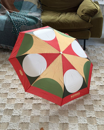 The Botero Patterned Umbrella - Red/Green/Yellow by Botero, featuring vibrant red, yellow, green, and white geometric designs, is open on a beige checkered rug beside a green armchair and a fringed blanket.