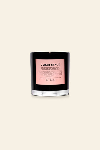 A black glass Boy Smells Cedar Stack Scented Candle 8.5oz jar with a pink label and description sits against a light beige background, highlighting the modern style of this signature candle from Boy Smells.