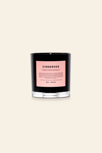 A black glass Boy Smells Cinderose Scented Candle 8.5oz with a pink label sits against a beige background. The unused candle, featuring an unlit wick, offers a delicate cinder rose floral fragrance.