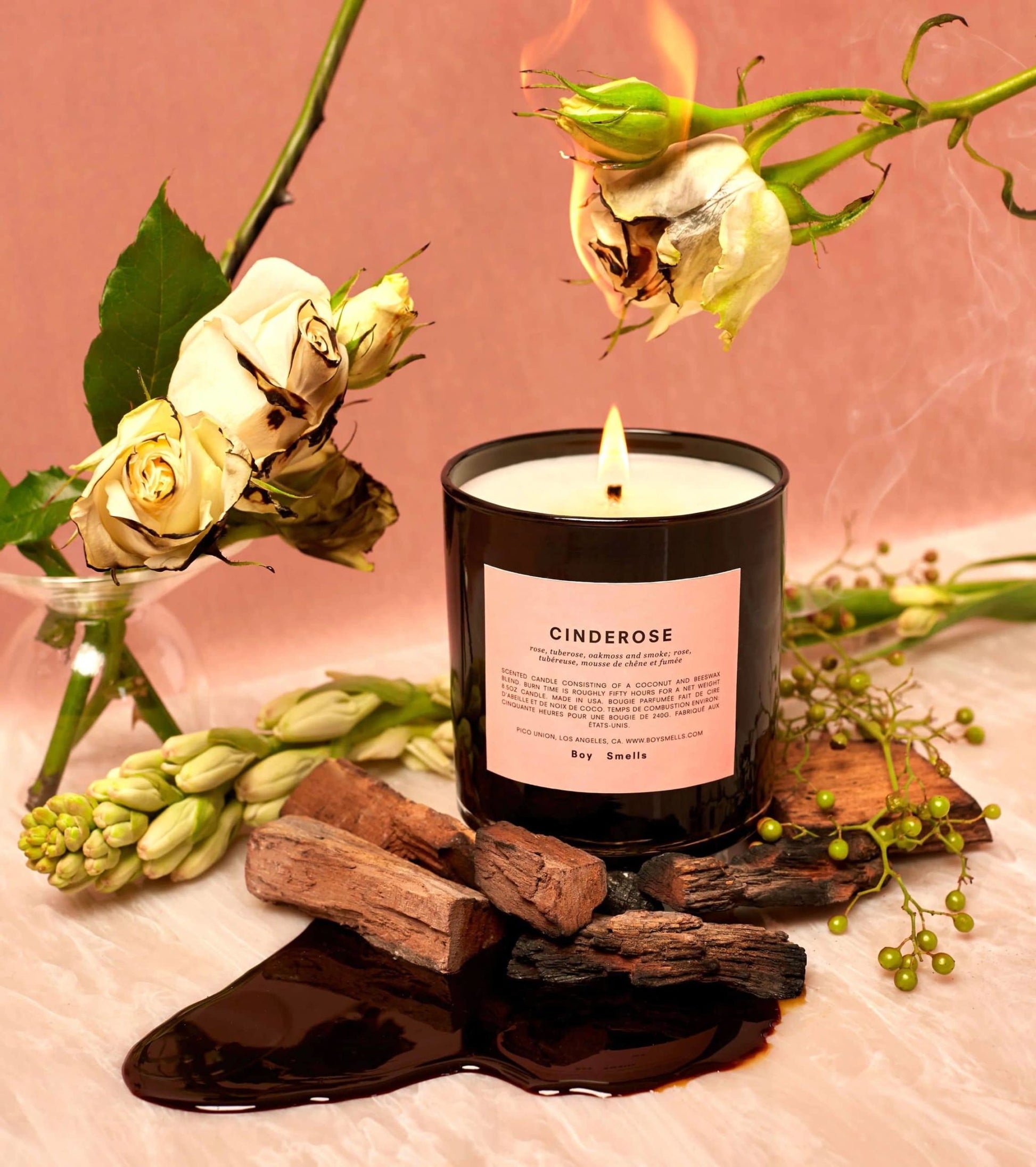 The Boy Smells Cinderose Scented Candle 8.5oz sits on marble among wilting white roses, dark liquid, wood pieces, green buds, and a burning rose—evoking its floral scent against a pink backdrop.