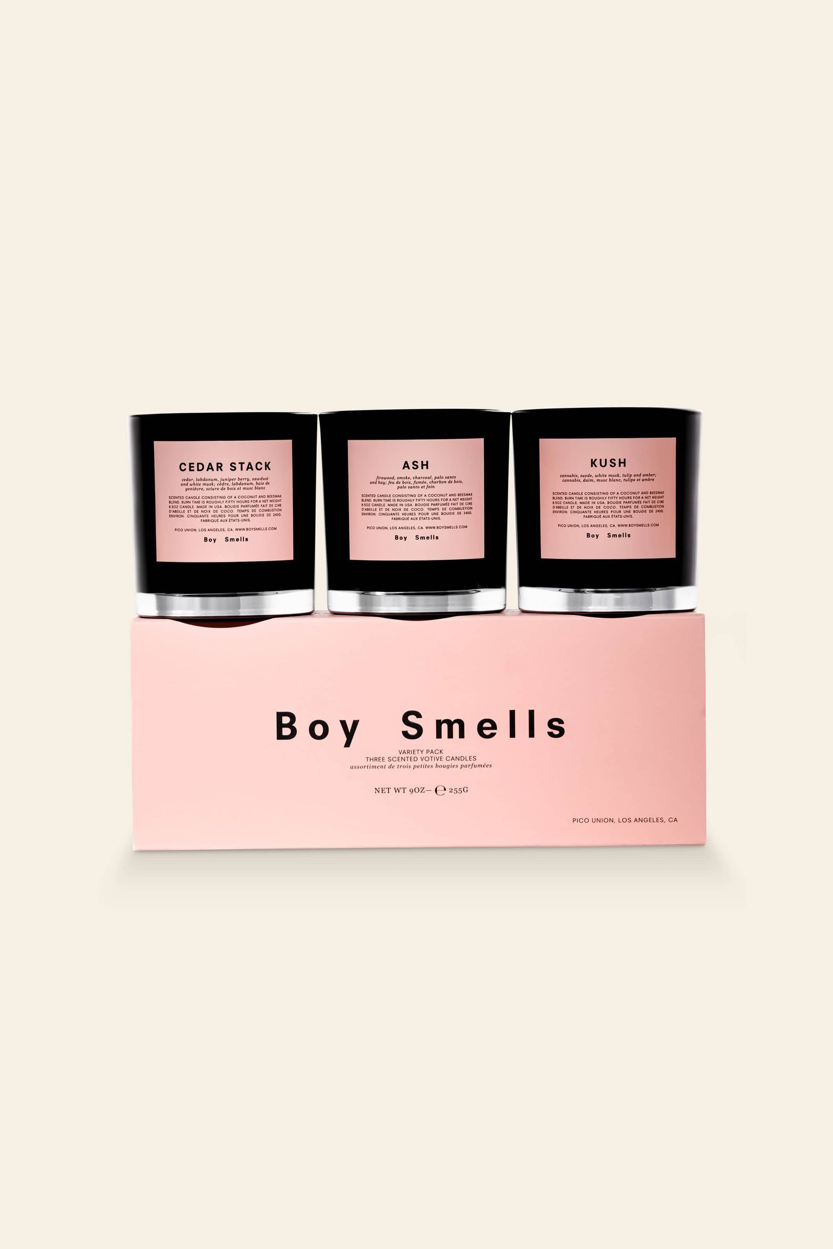 The Boy Smells Kush, Ash and Cedar Stack Votive Set features three petite candles with pink labels on a matching box, creating a chic look against a light beige background.