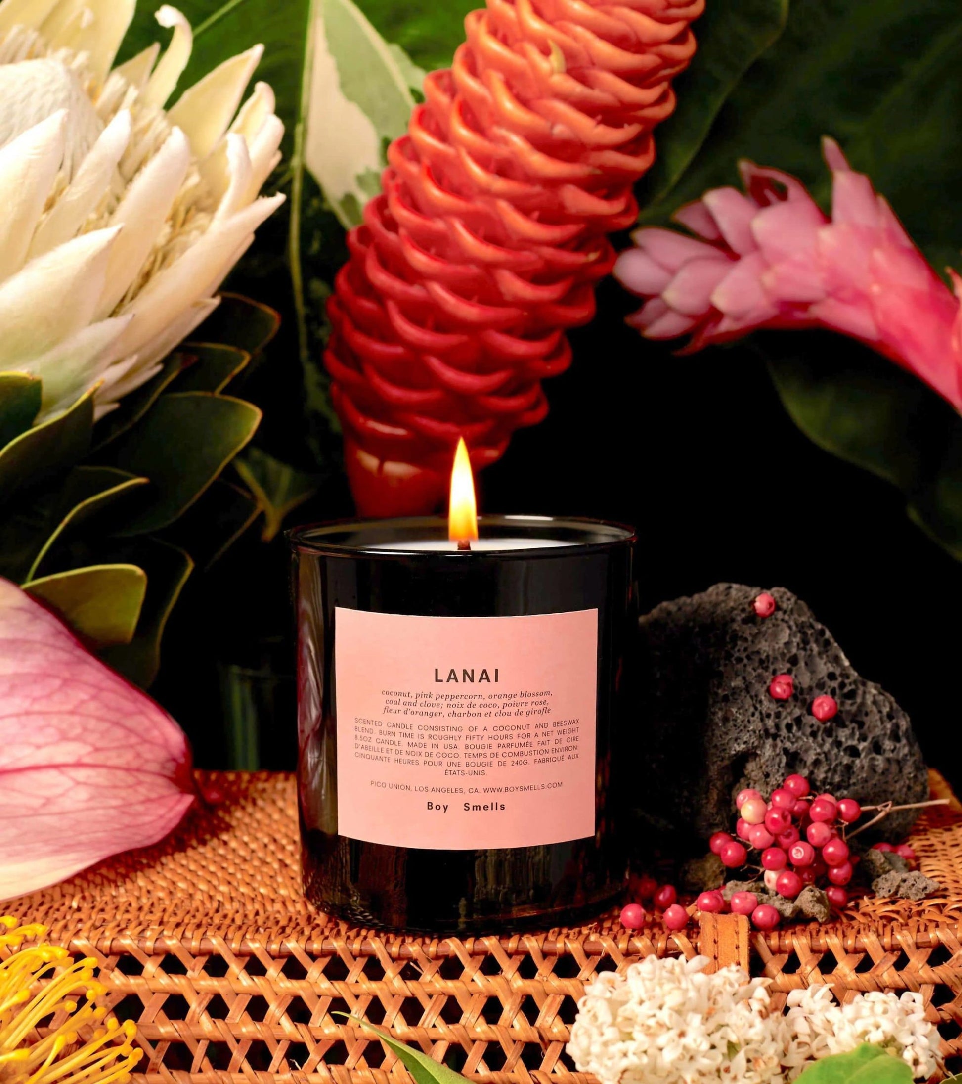 A lit Boy Smells Lanai Scented Candle 8.5oz sits on a woven surface, surrounded by coconut, tropical flowers, green leaves, volcanic rock, and pink peppercorns for an exotic luxury feel.