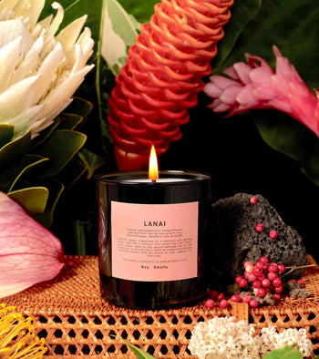 A lit Boy Smells Lanai Scented Candle 8.5oz sits on a woven surface, surrounded by coconut, tropical flowers, green leaves, volcanic rock, and pink peppercorns for an exotic luxury feel.