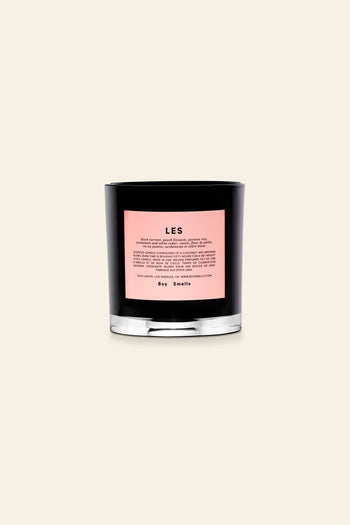 The Boy Smells Les Scented Candle 8.5oz features a black glass jar with a bold pink LES label and product details, plus Boy Smells branding at the bottom. Set against a beige background, it’s an ideal gift for lovers of unique fragrances.