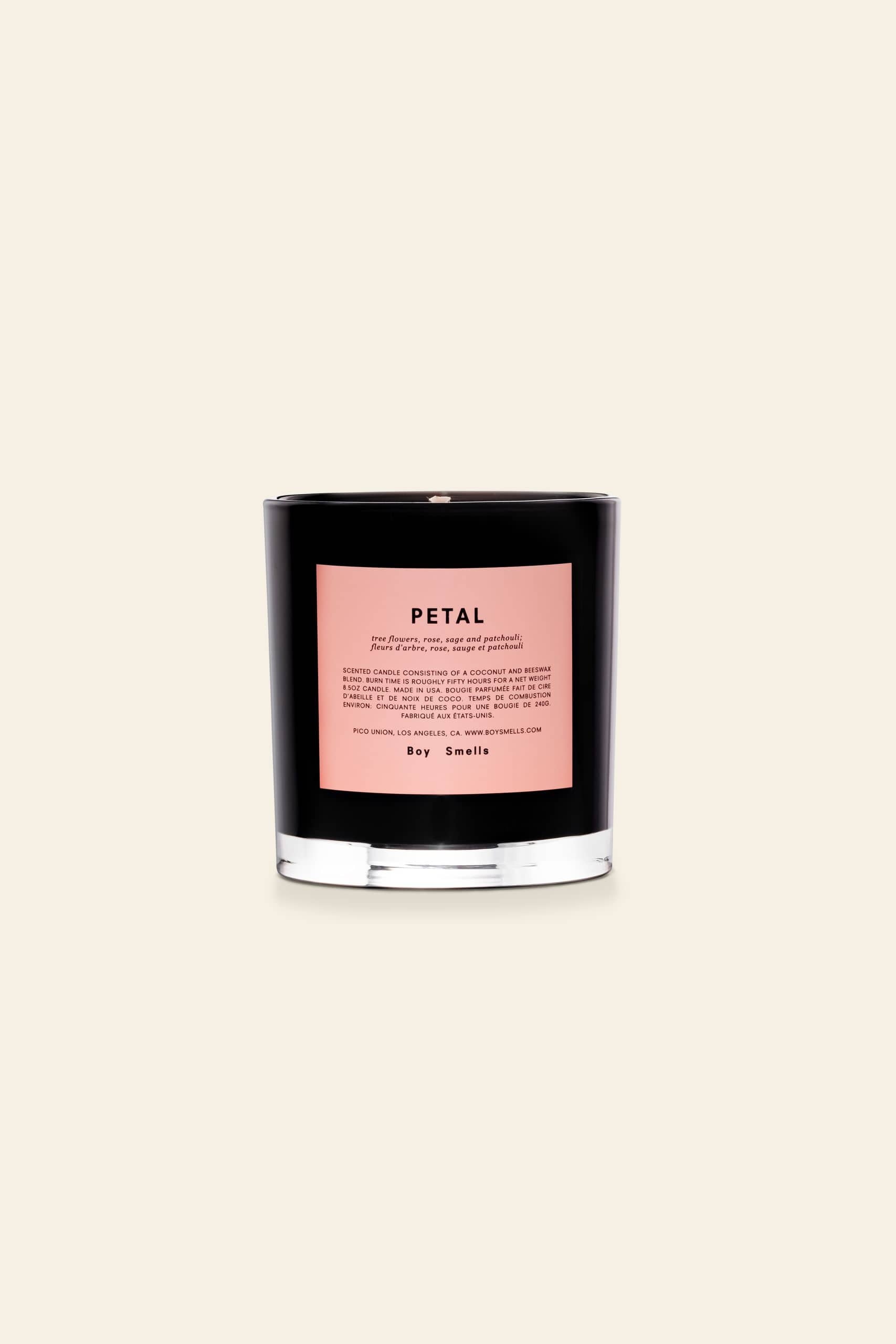 The Boy Smells Petal Scented Candle 8.5oz features a black glass vessel with a pink label and black text. It sits on a light beige background and offers an enchanting scent of exotic musk and magnolia.