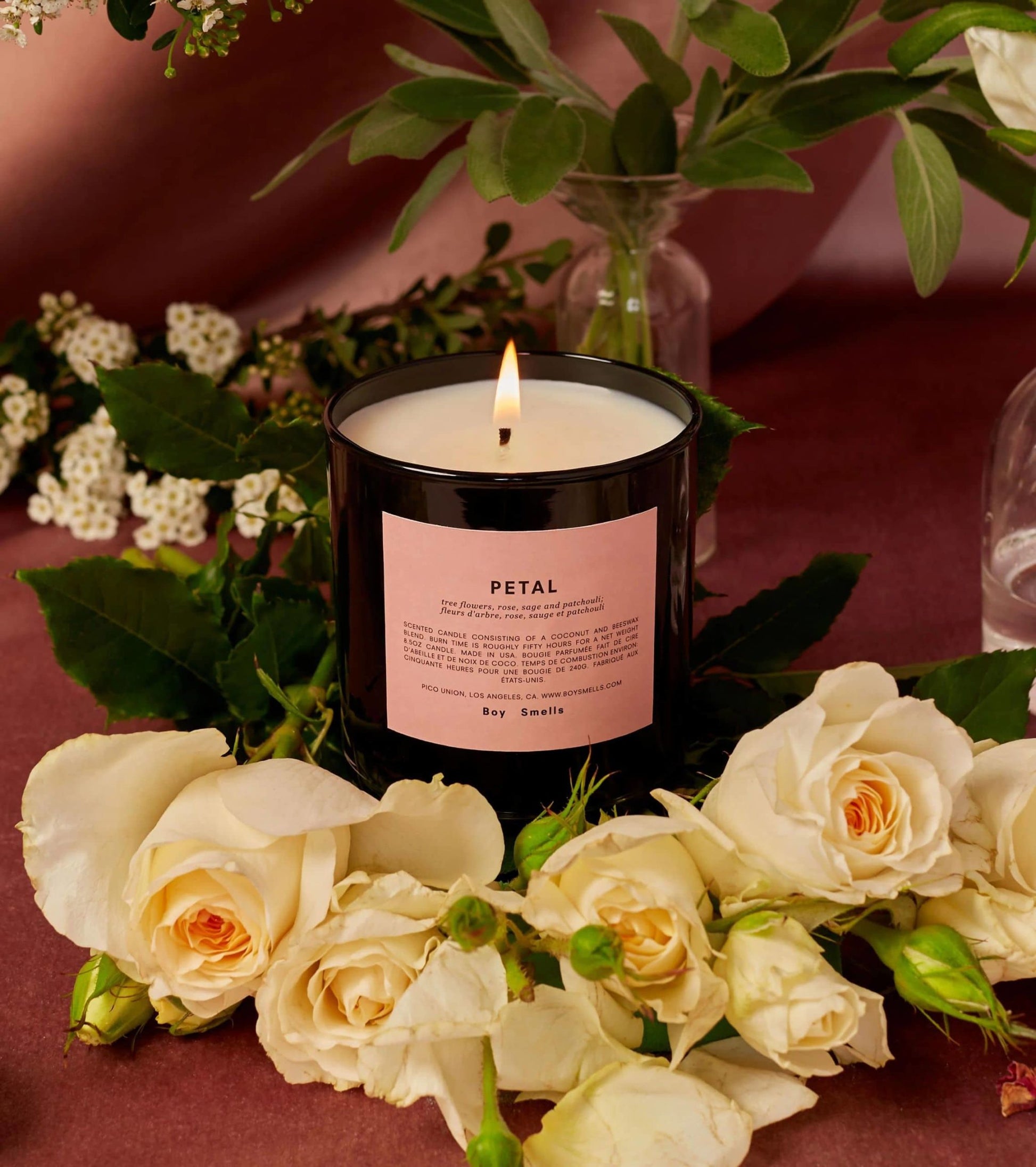 The Boy Smells Petal Scented Candle 8.5oz glows in a dark glass jar on a mauve surface, surrounded by white roses, greenery, and magnolia accents. A glass vase and flowers add to the tranquil setting. Brand: Boy Smells.