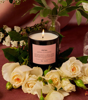 The Boy Smells Petal Scented Candle 8.5oz glows in a dark glass jar on a mauve surface, surrounded by white roses, greenery, and magnolia accents. A glass vase and flowers add to the tranquil setting. Brand: Boy Smells.