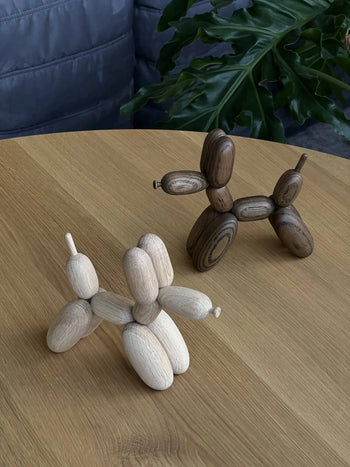 Two Boyhood Ballon Dog Small sculptures by Boyhood, crafted from European FSC certified oak in light and dark finishes, rest playfully on a light wood table with a green plant and part of a gray sofa in the background.