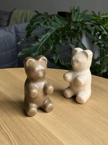 Two Boyhood Gummy Bear Small wooden figurines by Boyhood sit on a wooden table, with a leafy plant and gray couch in the background, creating a playful candy-inspired display.