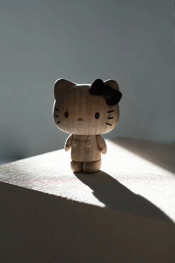 The Boyhood Hello Kitty Small by Boyhood features a wooden figurine with a black bow, standing on a sunlit surface and casting a long shadow—a charming collectible for any Hello Kitty or wooden figure enthusiast.