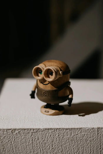 The Boyhood Minion Bob Small by Boyhood is a wooden figurine with round glasses and small black arms, posed playfully on a textured white surface under soft light, casting a shadow to the right.