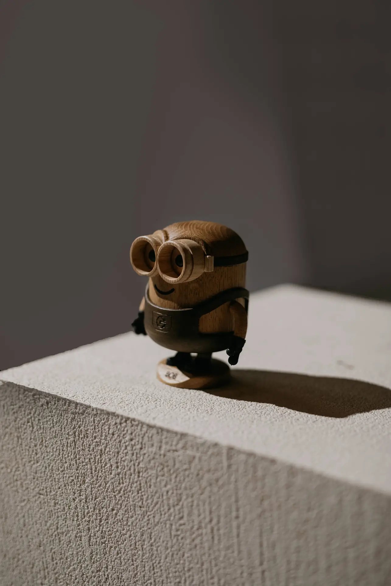 The Boyhood Minion Bob Small by Boyhood is a playful wooden figurine, standing on a textured light cube and casting a shadow against a softly lit, plain background.