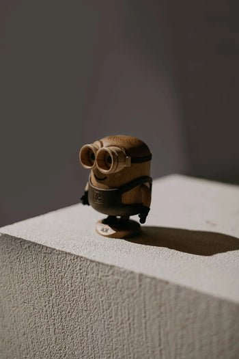 The Boyhood Minion Bob Small by Boyhood is a playful wooden figurine, standing on a textured light cube and casting a shadow against a softly lit, plain background.