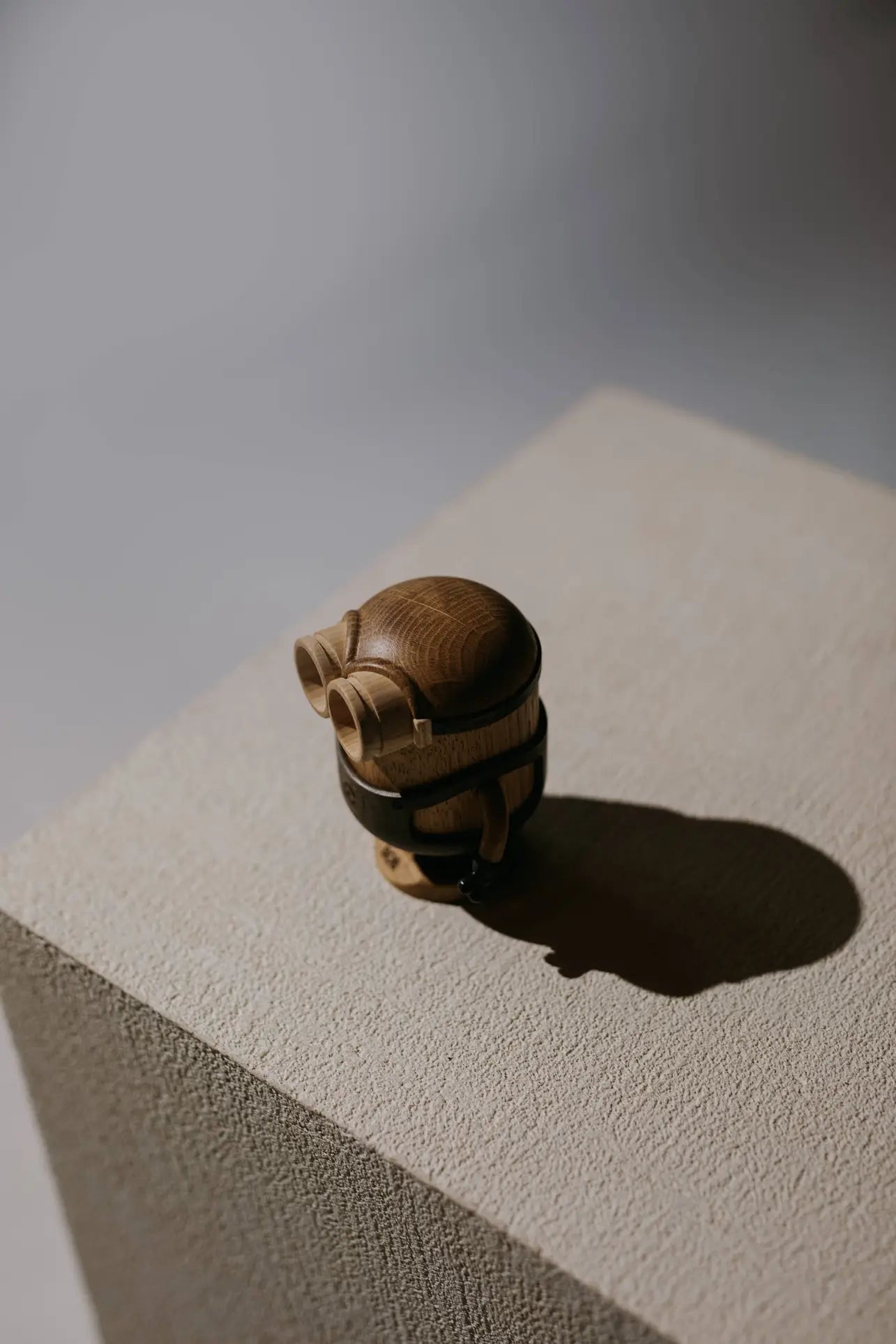 The Boyhood Minion Bob Small by Boyhood is a small, wooden, barrel-shaped figurine with goggles on a textured pedestal, playfully evoking the Minions movies as it casts a shadow.