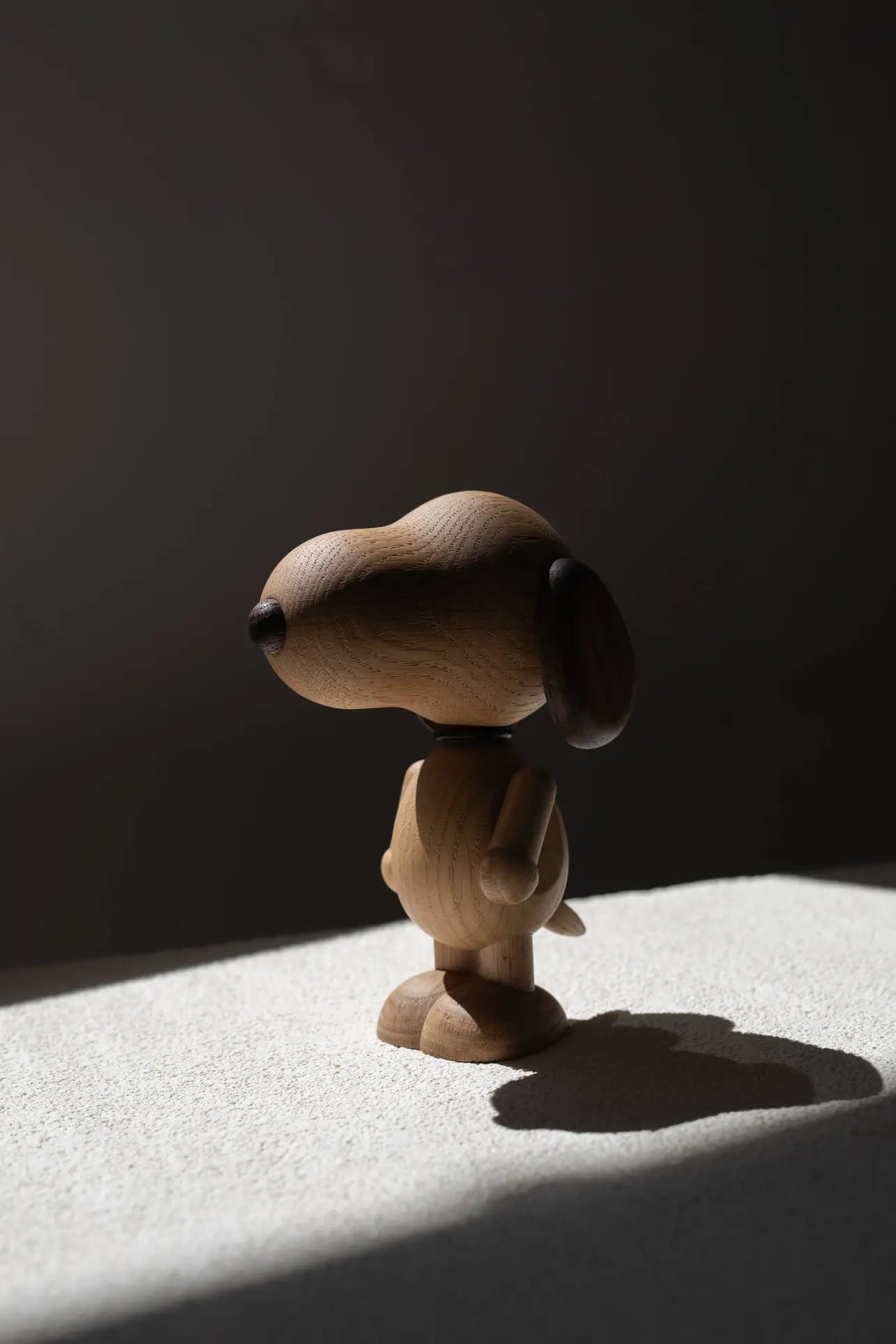 The Boyhood Snoopy Large figurine by Boyhood stands on a textured white surface, bathed in natural left light that casts a shadow rightward against a dark backdrop, evoking classic Charlie Brown scenes.
