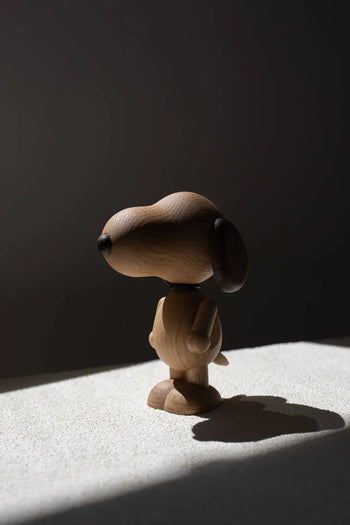The Boyhood Snoopy Large figurine by Boyhood stands on a textured white surface, bathed in natural left light that casts a shadow rightward against a dark backdrop, evoking classic Charlie Brown scenes.