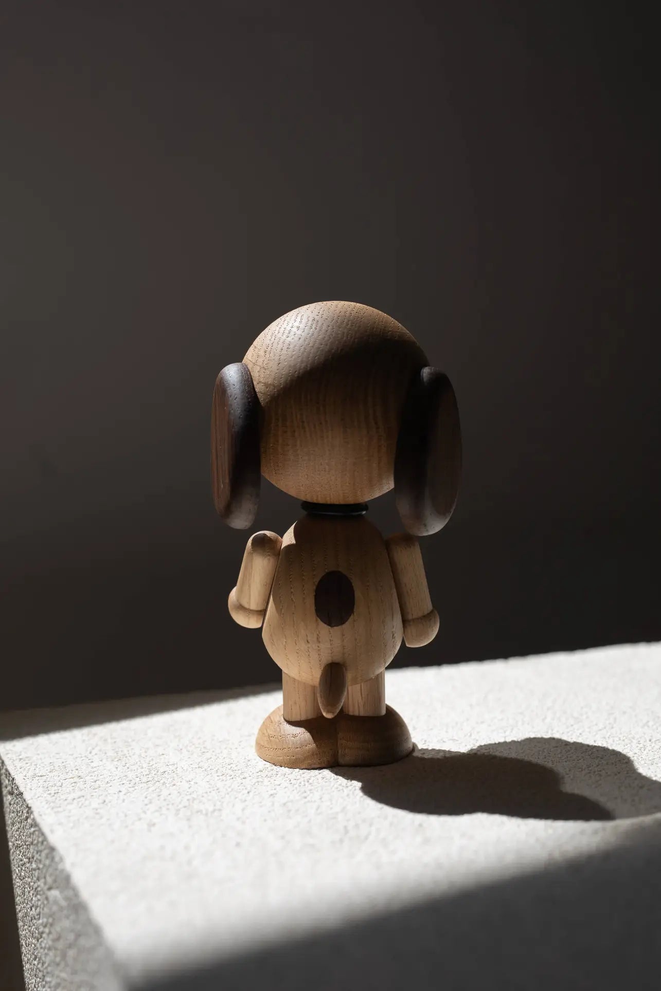 The Boyhood Snoopy Large by Boyhood is a wooden dog figurine with dark ears, standing on a textured surface and side-lit to cast a shadow against a dark background, reminiscent of the charm of Charlie Brown’s world.
