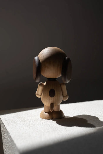 The Boyhood Snoopy Large by Boyhood is a wooden dog figurine with dark ears, standing on a textured surface and side-lit to cast a shadow against a dark background, reminiscent of the charm of Charlie Brown’s world.