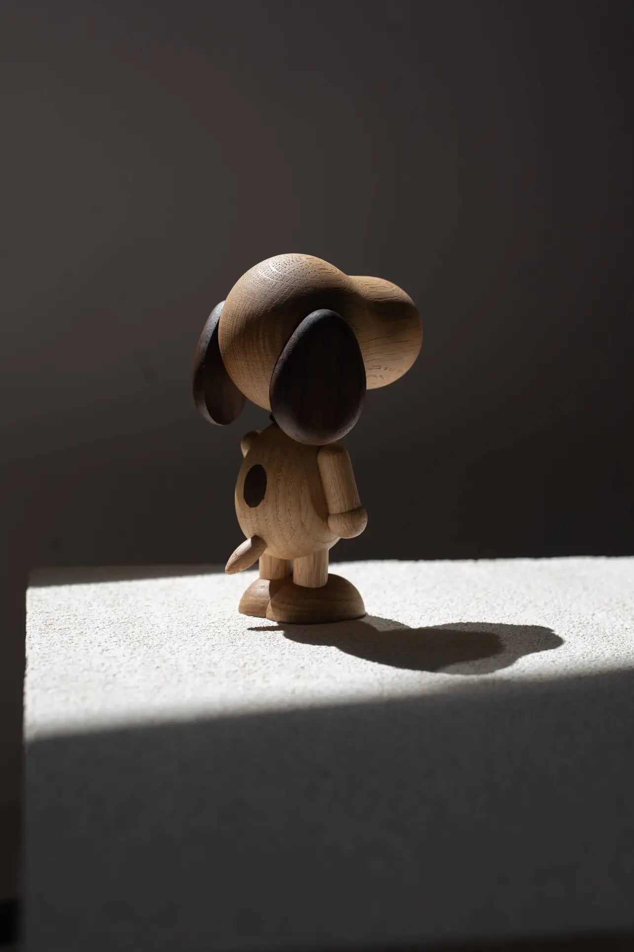 The Boyhood Snoopy Large by Boyhood, a sizeable wooden dog figurine inspired by Snoopy, stands on a light-colored surface with its shadow cast in soft, dramatic lighting.