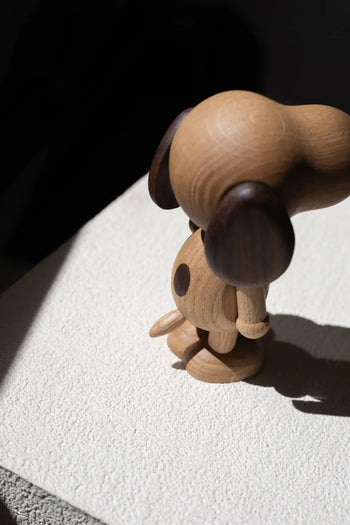 The Boyhood Snoopy Large by Boyhood, a wooden figurine featuring dark ears and a side spot, stands on a textured white surface bathed in sunlight, capturing the timeless charm of Charlie Brown’s iconic dog.