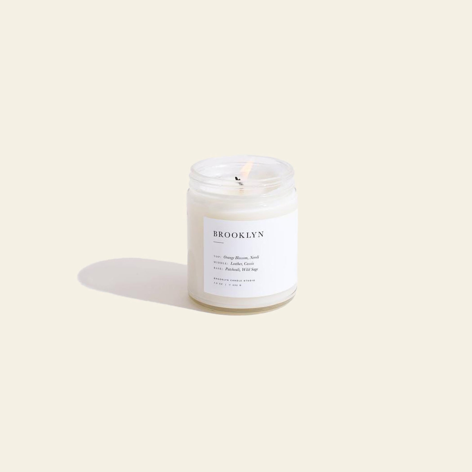 A Brooklyn Candle Studio Brooklyn Jar Candle in a clear glass jar rests on a light beige surface, casting a soft shadow. The label highlights notes of Orange Blossom and Sandalwood, evoking an elegant, timeless atmosphere with every scent.