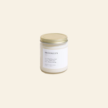 The Brooklyn Candle Studio Brooklyn Jar Candle comes in a glass jar with a gold lid and white label, featuring notes of Orange Blossom, Sandalwood, Lavender, Petitgrain, and Musk against a light beige background.