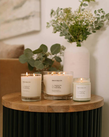 Three lit Brooklyn Candle Studio Brooklyn Jar Candles in glass jars are displayed on a round wooden table, with a white vase of green foliage and flowers in the background against a light wall.