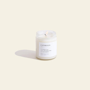 A lit Brooklyn Candle Studio Catskills Jar Candle glows in a clear glass jar on a light beige background, its label displaying woodsy fragrance notes and brand details, casting a soft shadow and evoking a calming ambiance.