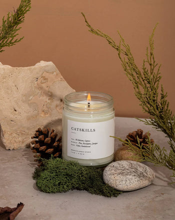 The Brooklyn Candle Studio Catskills Jar Candle burns atop a stone surface, spreading a woodsy scent. Moss, rocks, pinecones, and green sprigs surround it against beige and taupe stone for a calming ambiance.