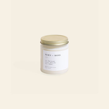 The Brooklyn Candle Studio Fern + Moss Jar Candle features a gold lid and rests on a light beige background. Its white label in minimal black text lists sage, lavender, and orange—nature-inspired notes crafted with essential oils.