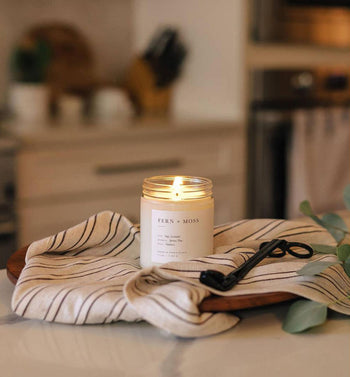 The Brooklyn Candle Studio Fern + Moss Jar Candle, known for its nature-inspired scent, is displayed on a striped cloth with a wick trimmer on a wooden tray atop a kitchen counter, with blurred kitchen items in the background.