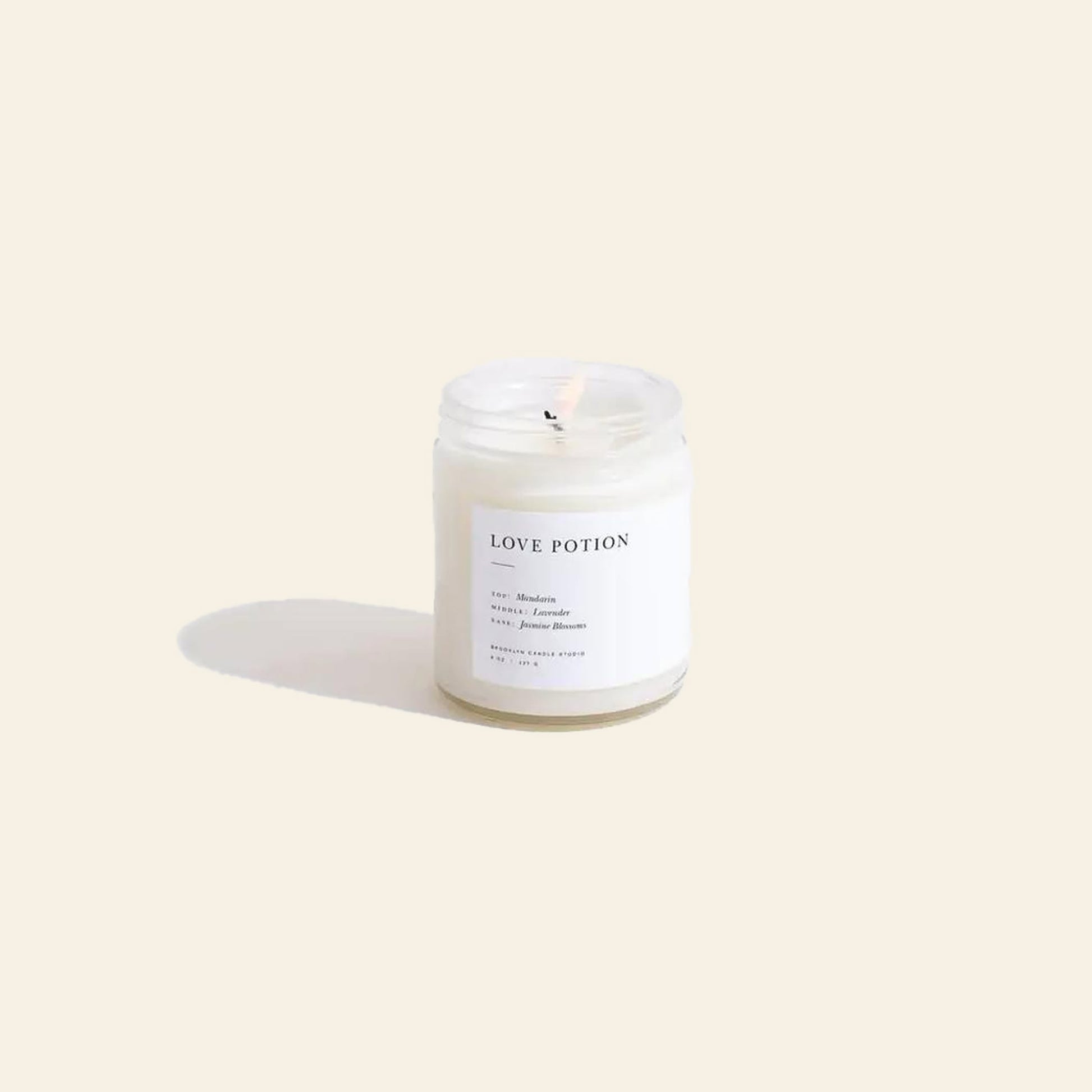 The Brooklyn Candle Studio Love Potion Jar Candle glows in a clear glass jar, casting gentle shadows on a beige surface as exotic jasmine blossoms fill the air with an enchanting aroma.