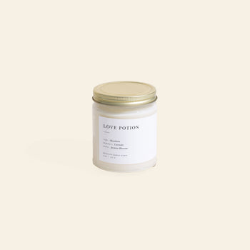 The Brooklyn Candle Studio Love Potion Jar Candle features notes of mandarin, jasmine blossoms, and dark amber in a glass jar with a gold lid. Its elegant label stands out against the plain light beige background.