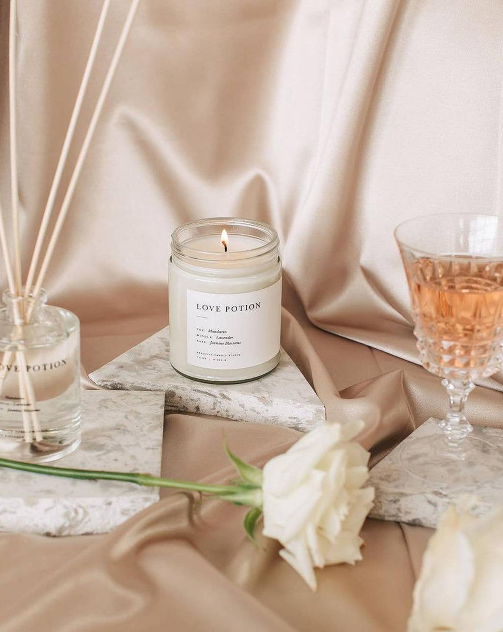 The Brooklyn Candle Studio Love Potion Jar Candle glows on marble beside a rose, rosé wine, and a reed diffuser atop beige satin, creating a soft, elegant, and romantic ambiance with its exotic jasmine aroma.