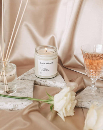 The Brooklyn Candle Studio Love Potion Jar Candle glows on marble beside a rose, rosé wine, and a reed diffuser atop beige satin, creating a soft, elegant, and romantic ambiance with its exotic jasmine aroma.
