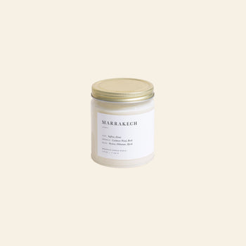 The Brooklyn Candle Studio Marrakech Jar Candle features a white glass jar with a gold lid and a minimalist label, infused with saffron and myrrh notes. It rests on a simple, light beige background.