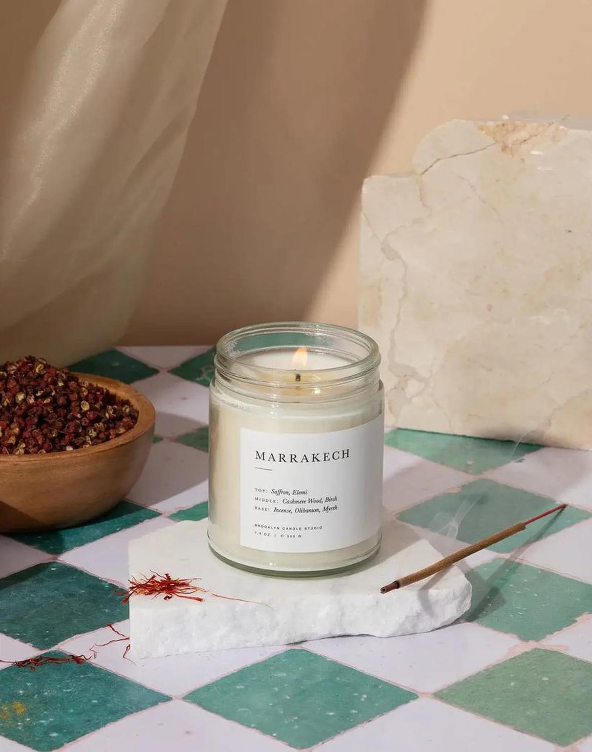 A lit Brooklyn Candle Studio Marrakech Jar Candle sits on a white stone slab beside a burning incense stick and a small bowl of spices, all arranged on a green-and-white checkered tiled surface.