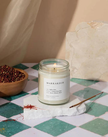 A lit Brooklyn Candle Studio Marrakech Jar Candle sits on a white stone slab beside a burning incense stick and a small bowl of spices, all arranged on a green-and-white checkered tiled surface.