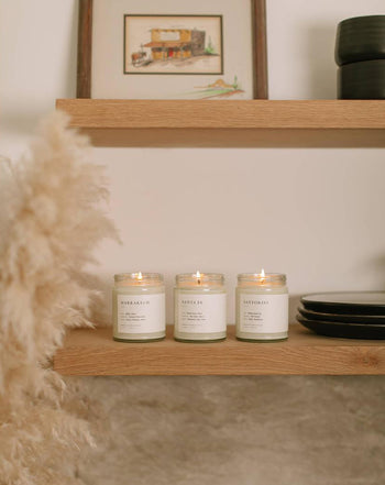 Three Brooklyn Candle Studio Marrakech Jar Candles in glass jars rest on a wooden shelf amid black plates, a cup, pampas grass, and art, filling the minimalist room with warm saffron and myrrh aromas.