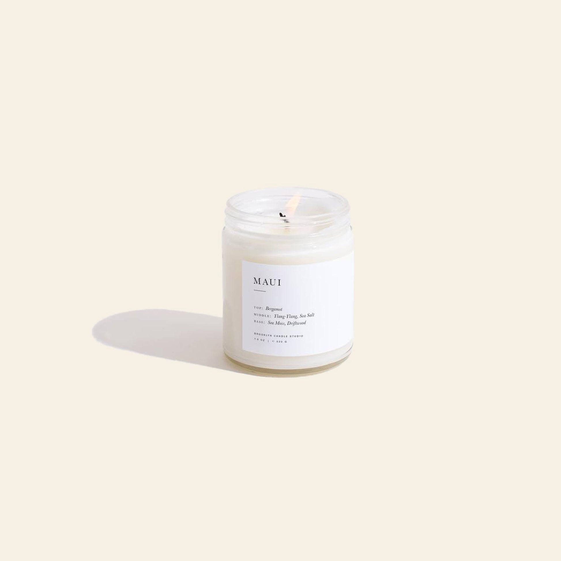 A glass Brooklyn Candle Studio Maui Jar Candle with a lit wick rests on a beige background, casting a soft shadow. The minimalist MAUI label lists fragrance notes, evoking an ocean-inspired atmosphere like the Hawaiian coast.