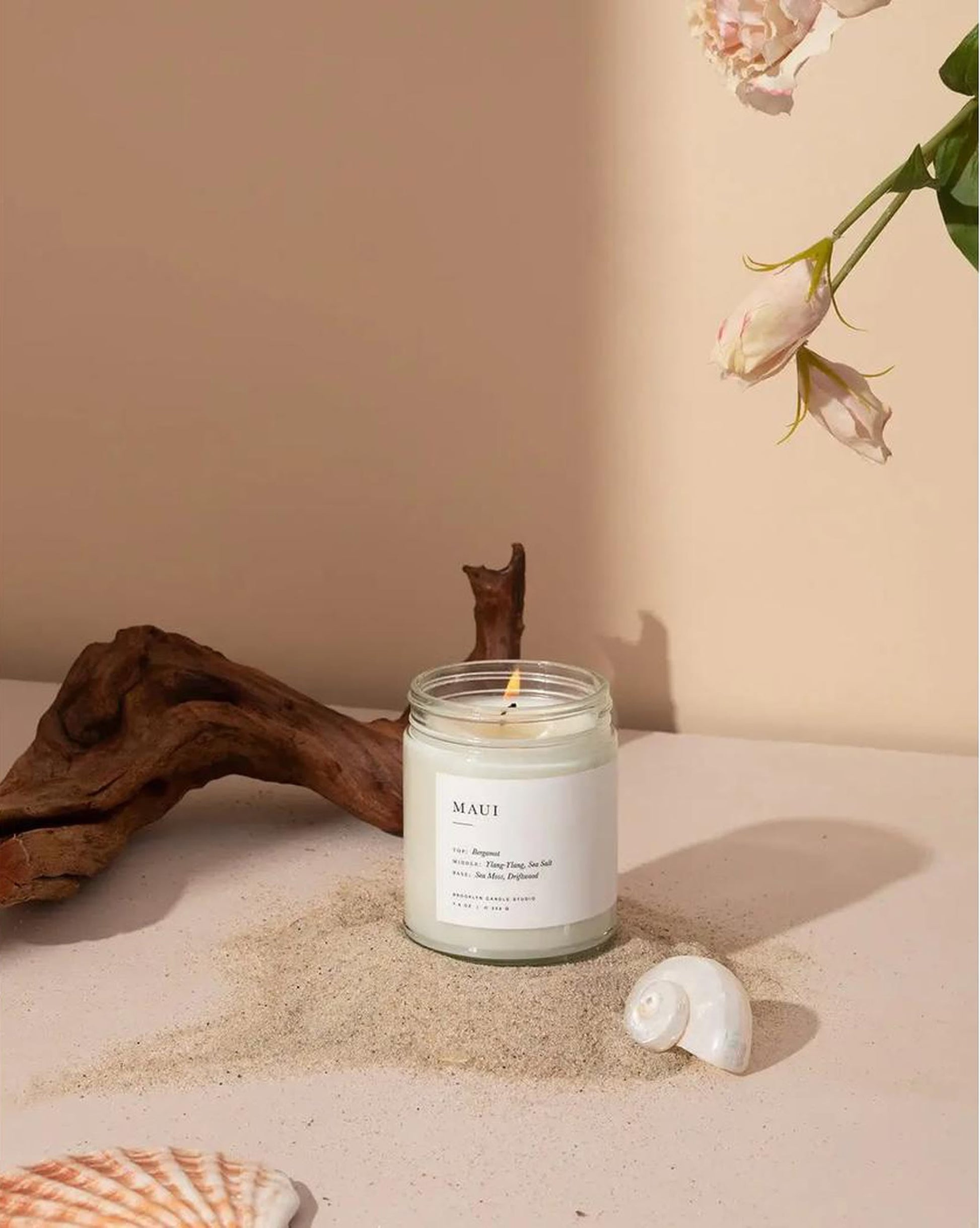 The Brooklyn Candle Studio Maui Jar Candle rests on sand beside seashells, driftwood, and pink flowers, creating an ocean-inspired Hawaiian vibe against a beige background.