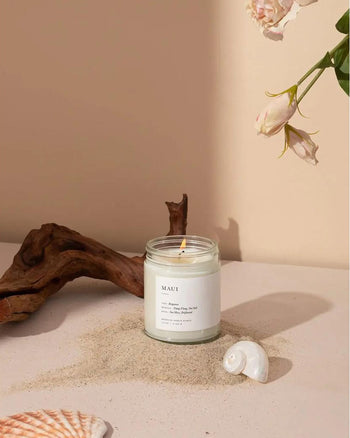 The Brooklyn Candle Studio Maui Jar Candle rests on sand beside seashells, driftwood, and pink flowers, creating an ocean-inspired Hawaiian vibe against a beige background.