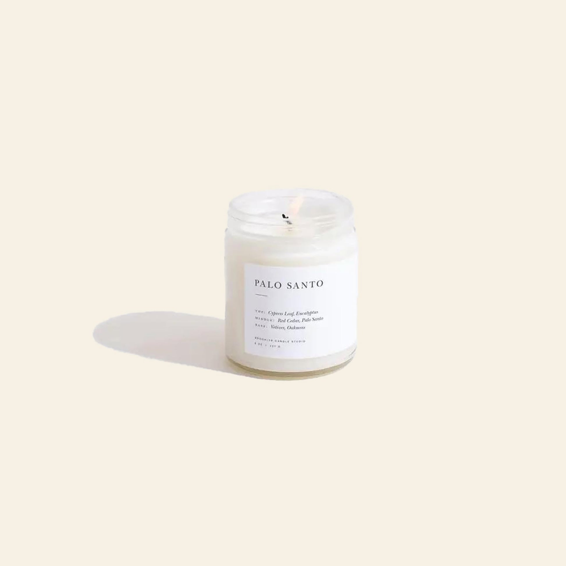 A lit Brooklyn Candle Studio Palo Santo Jar Candle in a clear glass jar rests on a beige background, its warm glow and soft shadow evoking the calming essence of cleansing rituals inspired by the sacred Palo Santo tree.