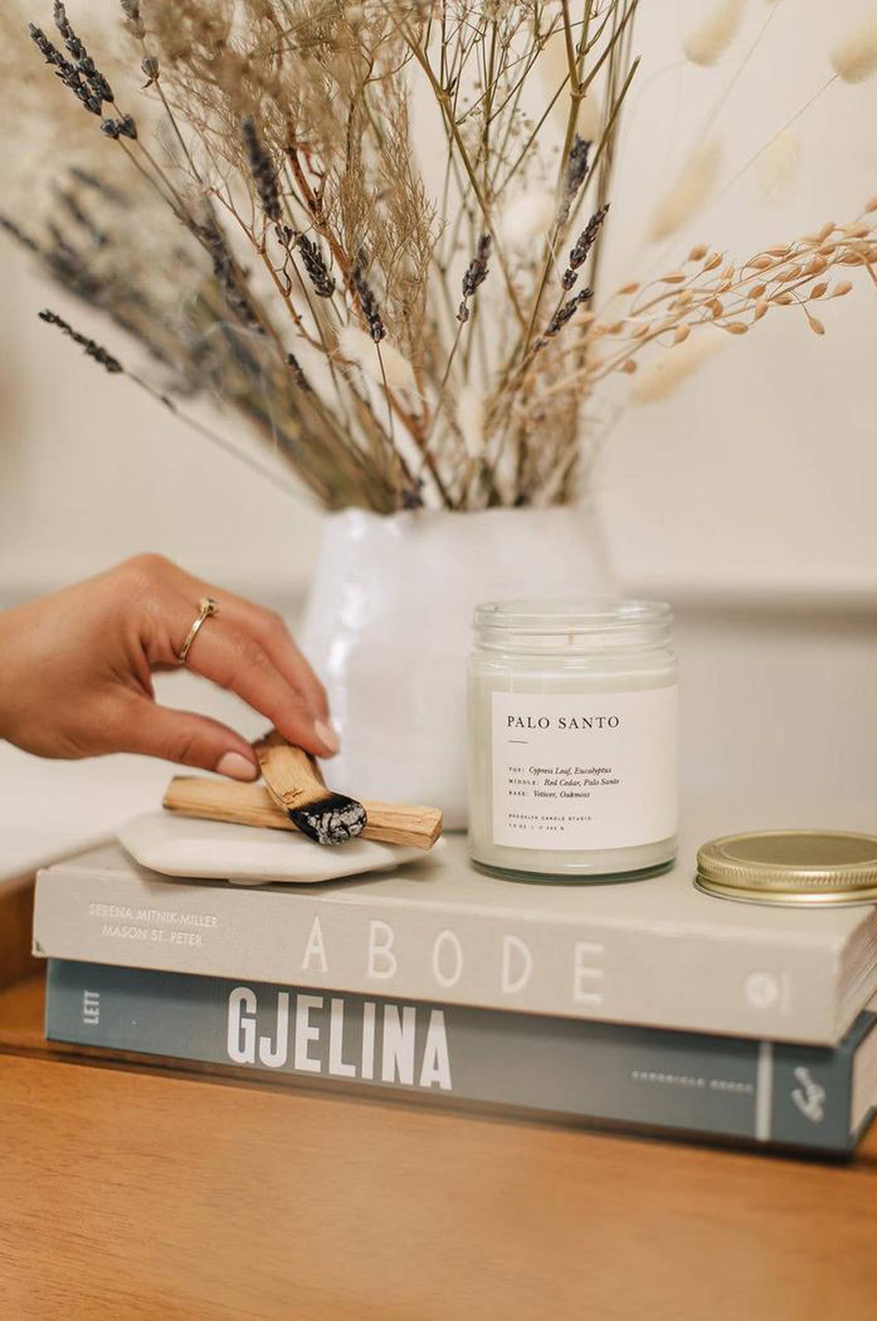 A hand lights wood from the sacred Palo Santo tree beside a Brooklyn Candle Studio Palo Santo Jar Candle on stacked books, with dried flowers in the background—ideal for cleansing rituals.