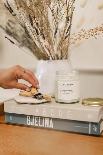 A hand lights wood from the sacred Palo Santo tree beside a Brooklyn Candle Studio Palo Santo Jar Candle on stacked books, with dried flowers in the background—ideal for cleansing rituals.