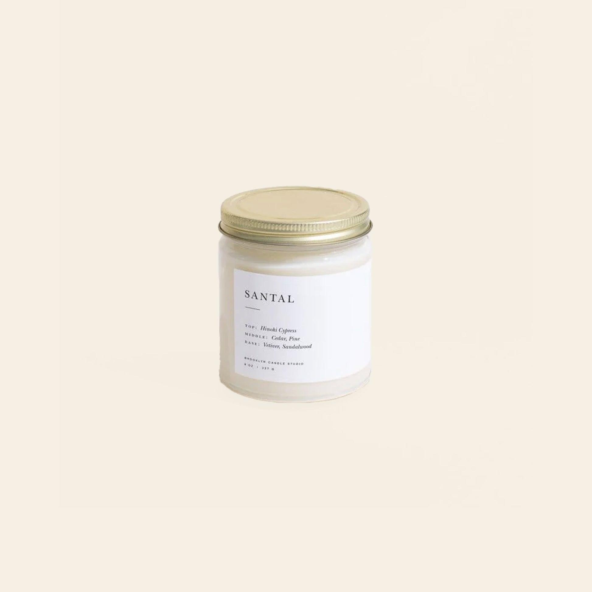 The Brooklyn Candle Studio Santal Jar Candle features a white glass jar with a gold lid, showcased on a light beige background. Its label highlights masculine, woodsy notes of hinoki cypress, cedar, and sandalwood.