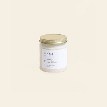 The Brooklyn Candle Studio Santal Jar Candle features a white glass jar with a gold lid, showcased on a light beige background. Its label highlights masculine, woodsy notes of hinoki cypress, cedar, and sandalwood.