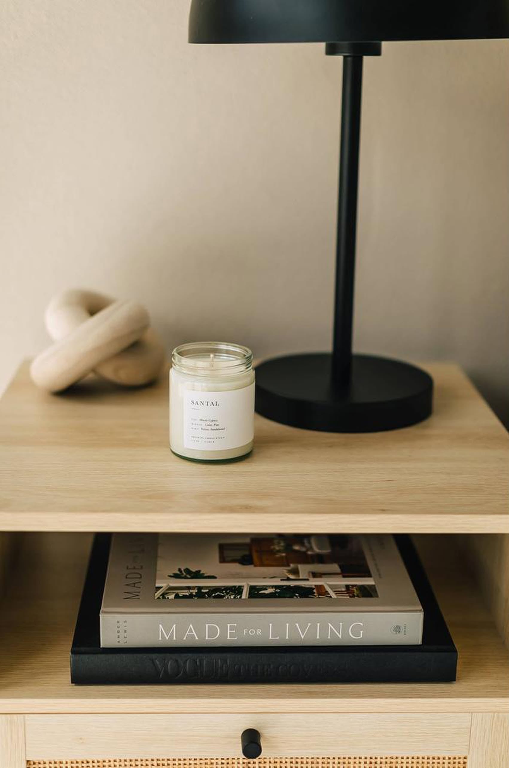 A wooden nightstand holds a black lamp, a decorative object, and the Brooklyn Candle Studio Santal Jar Candle, featuring a masculine, woodsy scent. On the lower shelf are two books: MADE FOR LIVING and VOGUE.